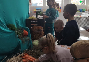 Dzieci oglądają leżące na sianie warzywa. Dotykają produktów, są zaciekawione.